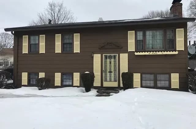 A brown, two-story house with cream shutters and a metal front door stands amidst a snowy yard. Bare trees and a chimney hint at a cold, winter day.