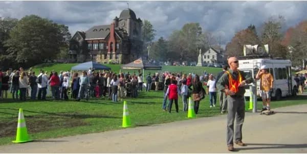A crowd gathers on a lawn in front of a large, historic building under a cloudy sky. A person wearing a reflective vest is in the foreground, next to a van.