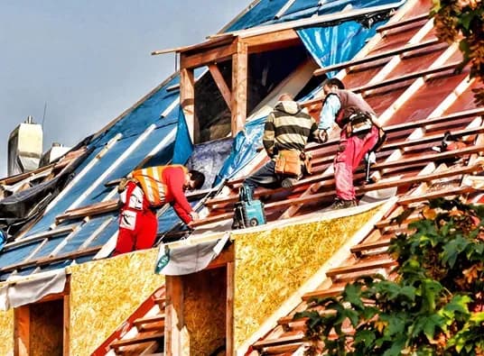 Three workers in safety gear repair a steep, colorful roof under a cloudy sky. They use tools and materials, conveying teamwork and determination.