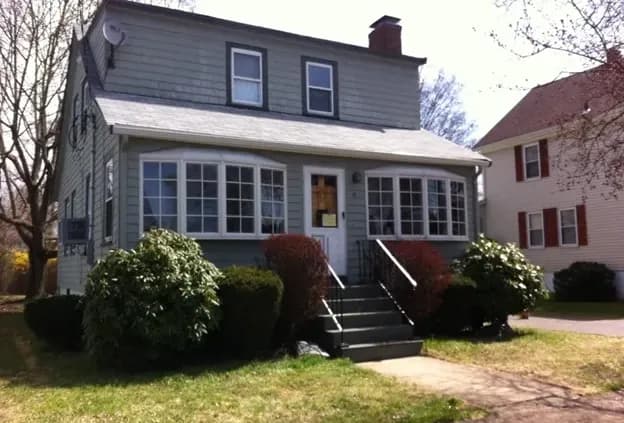 Two-story gray house with a gabled roof and front porch, adorned by green shrubs. Sunny day, neighboring house visible, conveying a serene suburban setting.