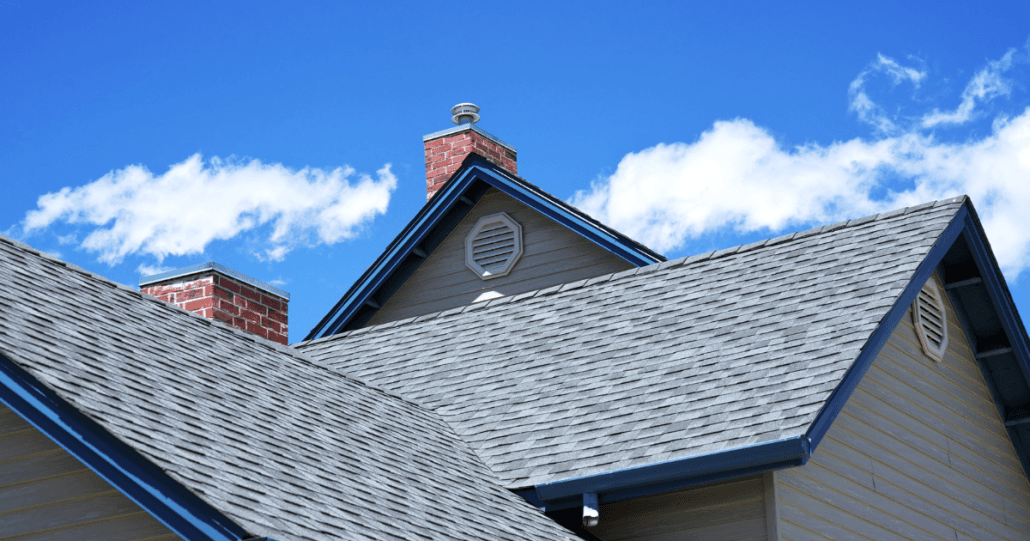 Gabled roof with gray shingles and red brick chimney against a bright blue sky with scattered clouds, conveying a serene and clear day.