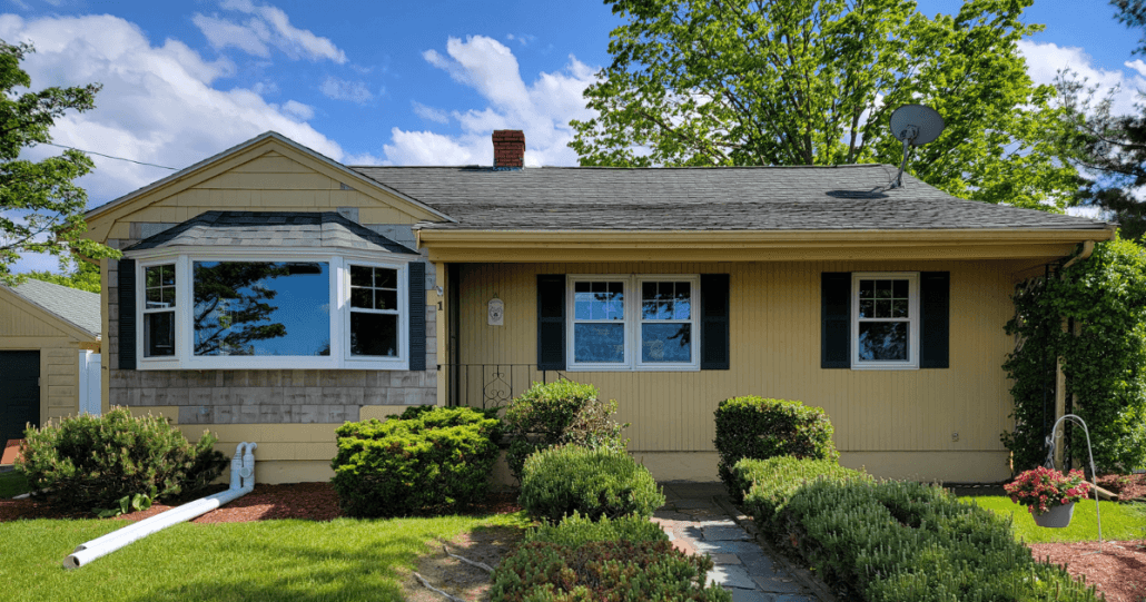 Single-story yellow house with dark shutters and a large bay window. Lush bushes, trimmed hedges, and a hanging flower pot in front yard. Sunny day, blue sky.