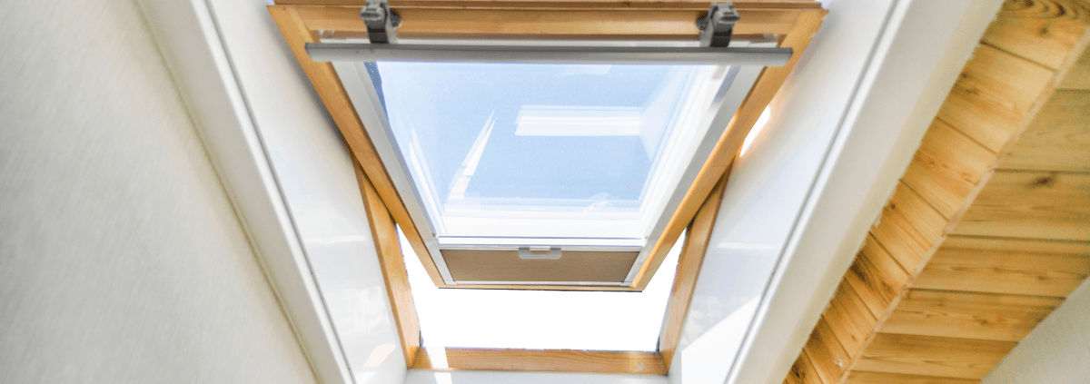 A wooden-framed skylight window partially open, letting in bright daylight. The surrounding ceiling is white, and wooden stairs are visible on the right.