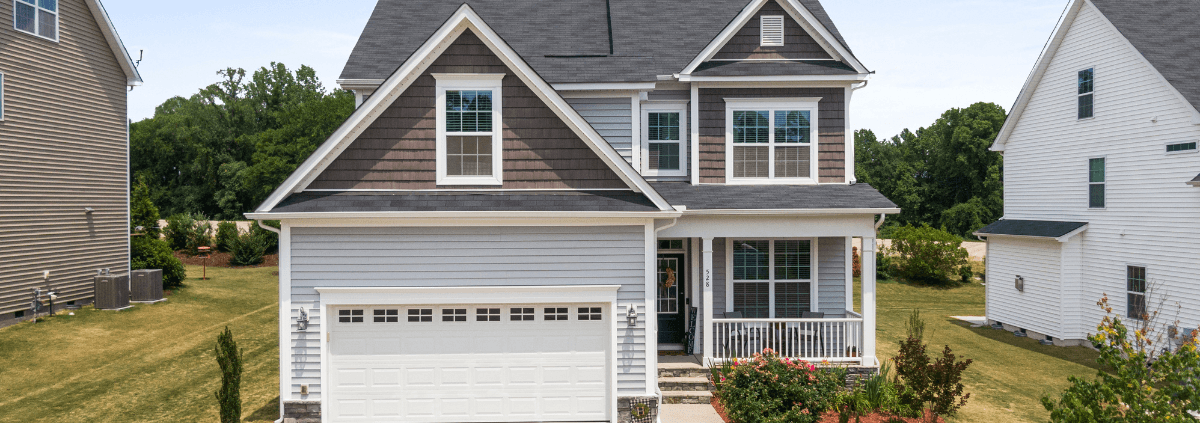 Two-story suburban house with a gabled roof, white and brown siding, and a spacious front porch. The well-tended lawn and vibrant bushes add charm.