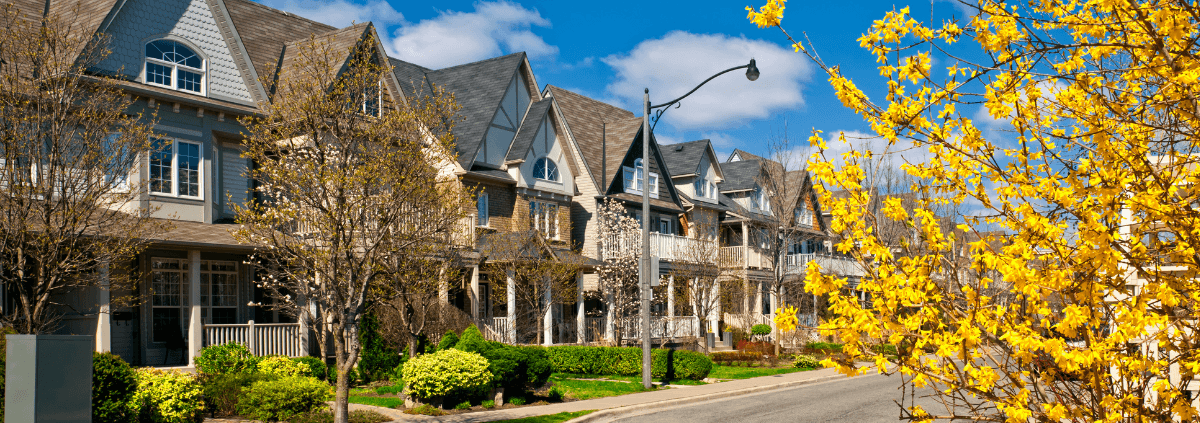 Row of charming suburban houses under a bright blue sky, surrounded by budding trees and vibrant yellow flowers, evoking a cheerful spring mood.