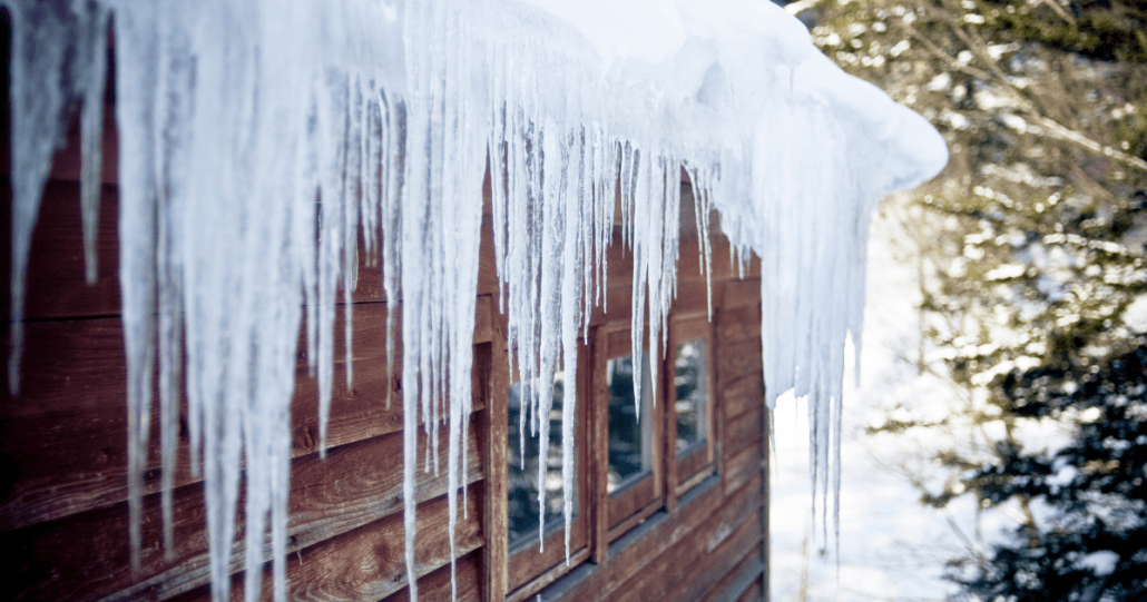 Icicles hang from a snowy roof of a wooden cabin in a winter forest. Sunlight glints off the ice, creating a serene and chilly atmosphere.