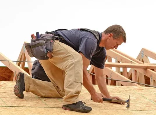A construction worker kneels on a wooden roof frame, hammering nails with focus. He wears a tool belt, blue shirt, and tan pants. The scene conveys determination.