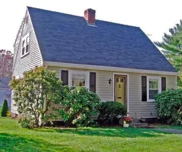 A cozy beige house with a dark roof, white trim, and a central chimney. Surrounded by lush shrubs and a well-kept lawn, it exudes a welcoming vibe.