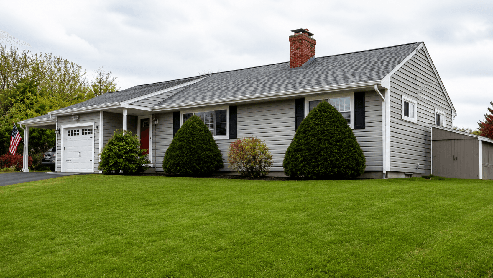 A single-story, light gray house with a chimney, and a sloped roof. It's surrounded by a green lawn and bushes, under a cloudy sky.
