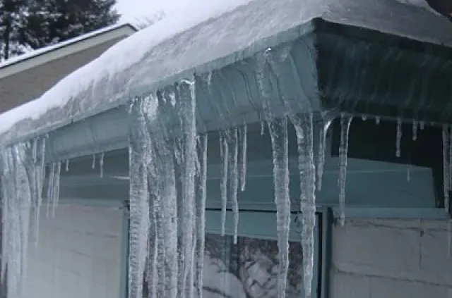 Icicles hang from a snowy roof edge, glistening in the cold winter light. The scene conveys a chilly, serene atmosphere with snow-covered surroundings.