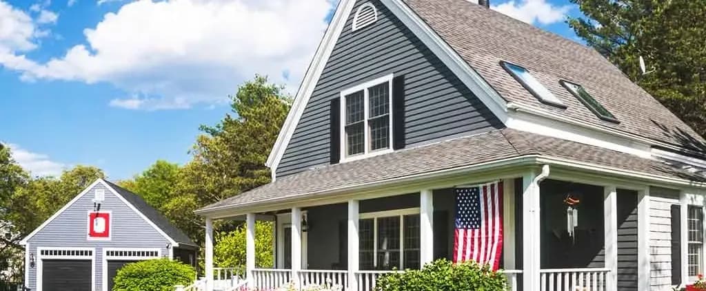 A charming gray house with a steep roof and dormer windows, displaying an American flag on the porch. A detached garage stands nearby, both surrounded by lush greenery under a bright blue sky.