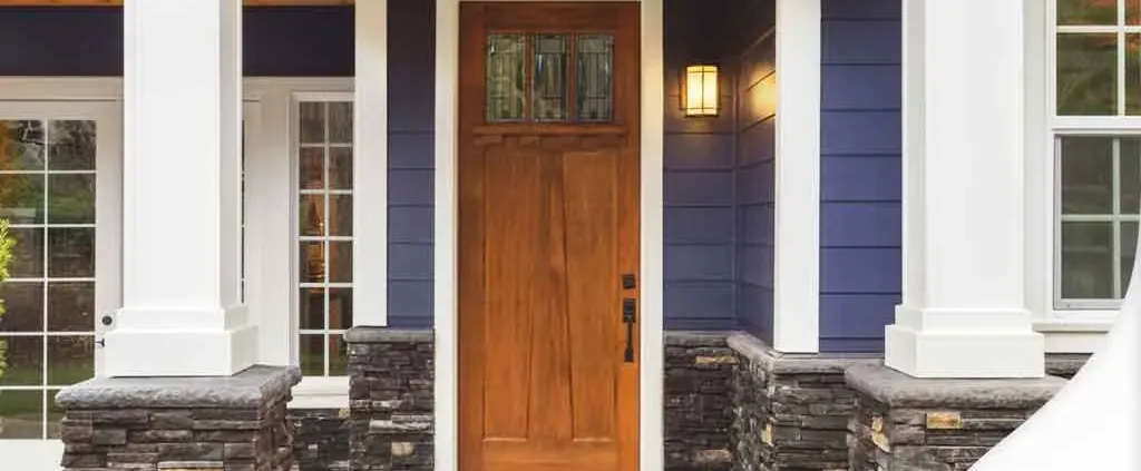 The image shows a welcoming front entrance with a wooden door, flanked by white columns and stone accents. The blue siding adds a modern touch.