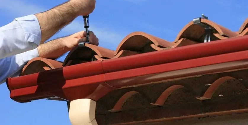 Hands fastening clay roof tiles with screws under a clear blue sky. The scene conveys precision and care in roofing maintenance.
