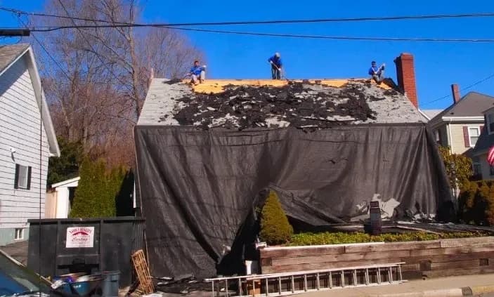 Three workers stand on a roof under repair, covered in black debris. A tarp hangs down the front, with a dumpster and ladder near the house. Clear sky above.