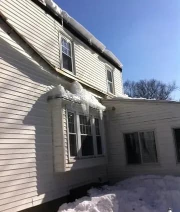 A two-story white house with icicles hanging from the roof, casting shadows on the snow. Clear blue sky in the background, evoking a chilly winter day.