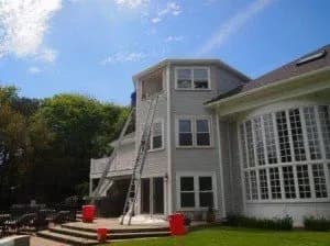 A large, three-story gray house with white trim under a clear blue sky. Ladders lean against it, with red buckets on a sunny patio, suggesting ongoing maintenance or repairs.