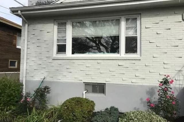 A white brick house wall with a large window at the center, surrounded by neatly trimmed bushes and flowering plants in a small garden bed below.