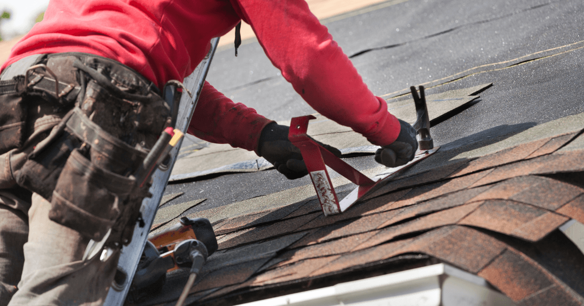 Worker in a red shirt repairs a shingle roof, using tools while balancing on a ladder. The scene conveys focus and precision in construction work.