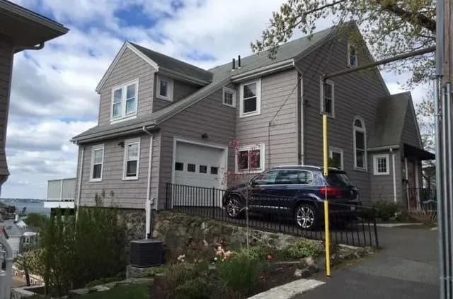 A two-story gray house with a sloped roof and white trim sits on a slight hill. A blue SUV is parked in the driveway. The sky is partly cloudy.