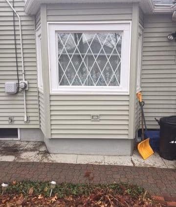 A house with gray siding features a bay window with diamond-patterned glass. Outside, a yellow snow shovel leans against the wall, next to a black trash can.