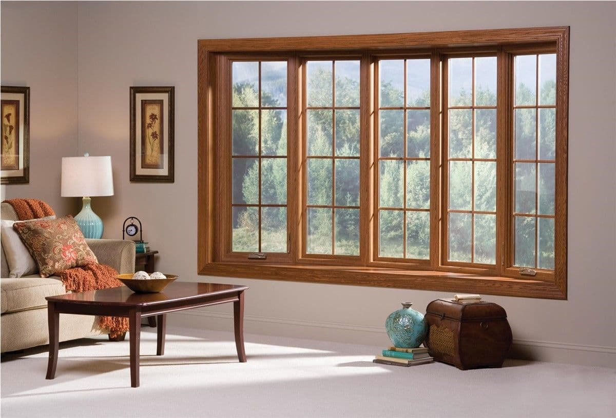 Cozy living room with a large bay window showcasing a forest view. Features a beige sofa, decorative pillows, wooden coffee table, and a lamp.