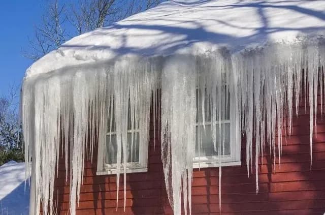 Red house with large icicles hanging from snow-covered roof against a clear blue sky. The scene conveys a cold, wintry atmosphere.