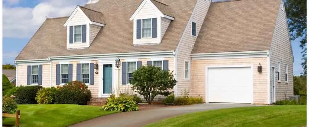 A charming Cape Cod-style house with beige siding, steep gabled roof, and two dormer windows. It features a blue front door, neat shrubs, and a lush green lawn.