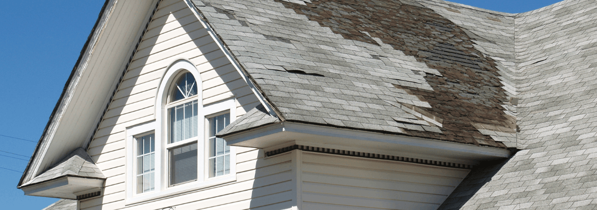 A house with a steep, shingle roof shows significant damage and missing tiles, contrasting with the clear blue sky. The tone is concerning and in need of repair.
