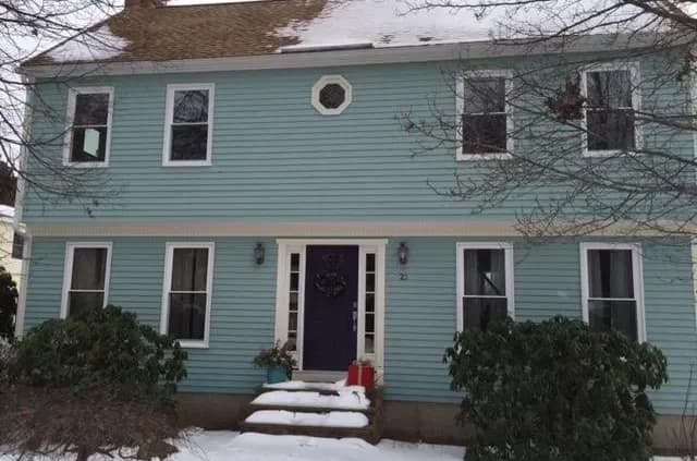 Two-story blue house with a gable roof and snow-covered steps. The front door is purple with a wreath, flanked by shrubs and bare trees.