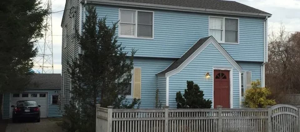 Two-story blue house with a red door, large windows, and a wooden fence. A tree and bushes are in the front yard; the tone is calm and residential.