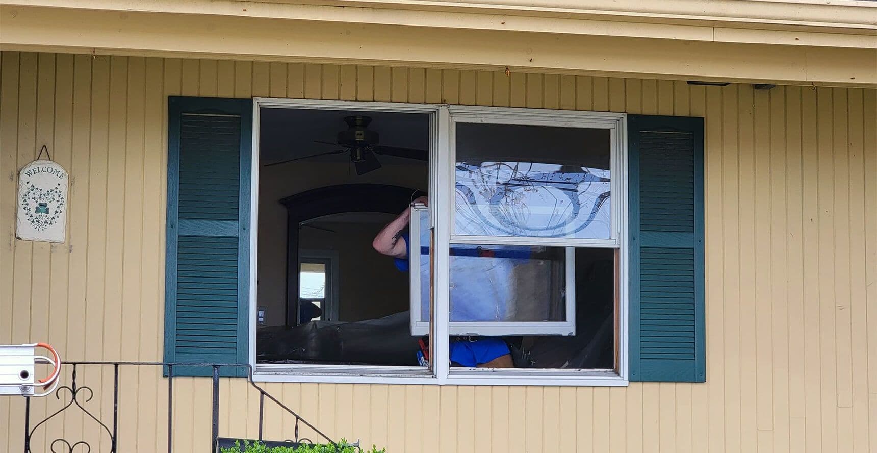 Person installing an air conditioner through an open window of a beige house with green shutters. A "Welcome" sign hangs on the wall, evoking a warm, inviting atmosphere.