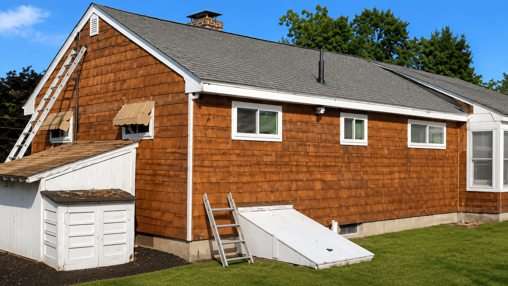 A wooden house with exposed walls during renovation. Blue tarp covers debris on the grass. A ladder leans against the house under a sloped roof.