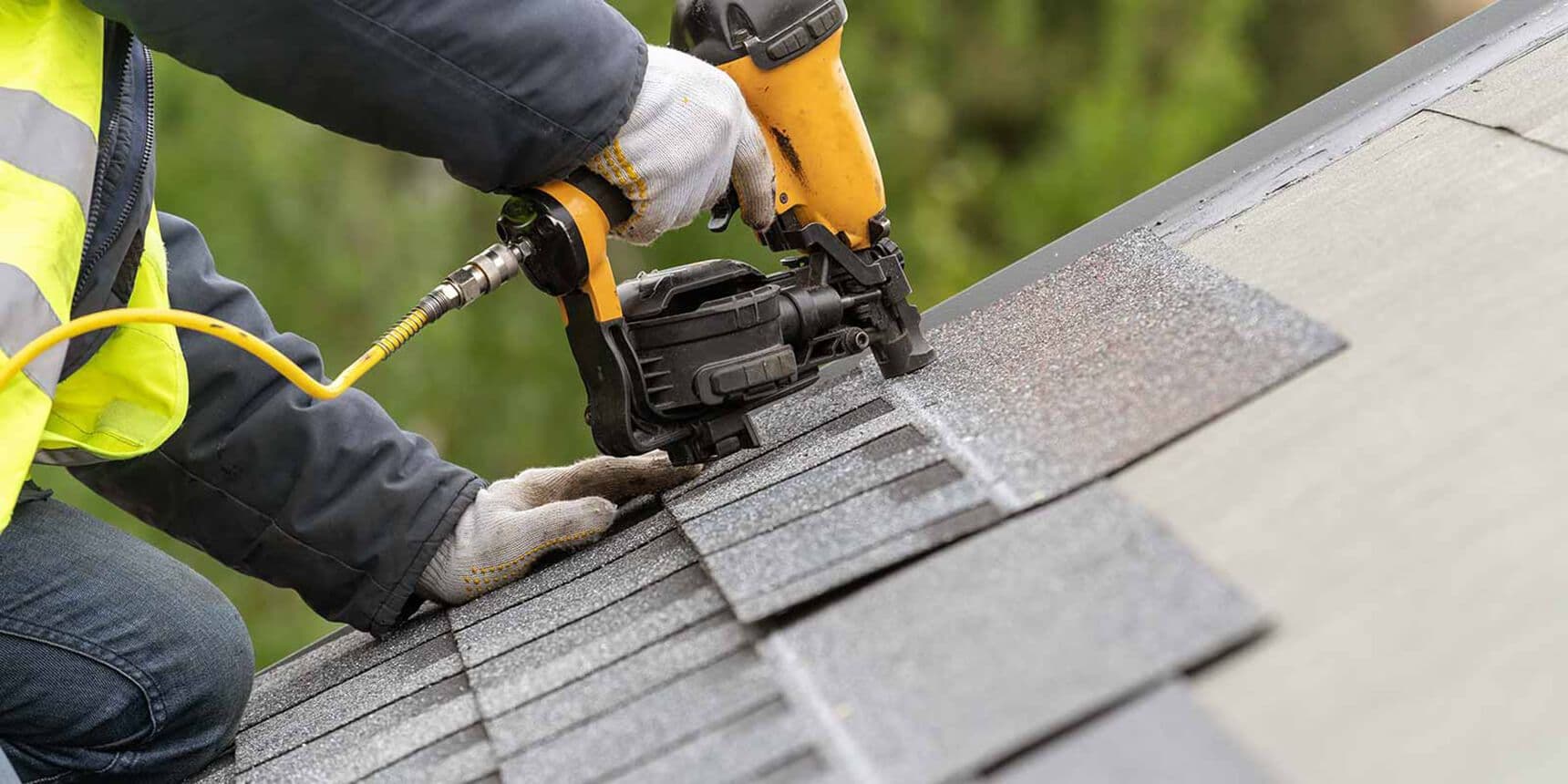A worker in safety gear uses a pneumatic nail gun to secure asphalt shingles on a roof. The scene conveys construction and precision.