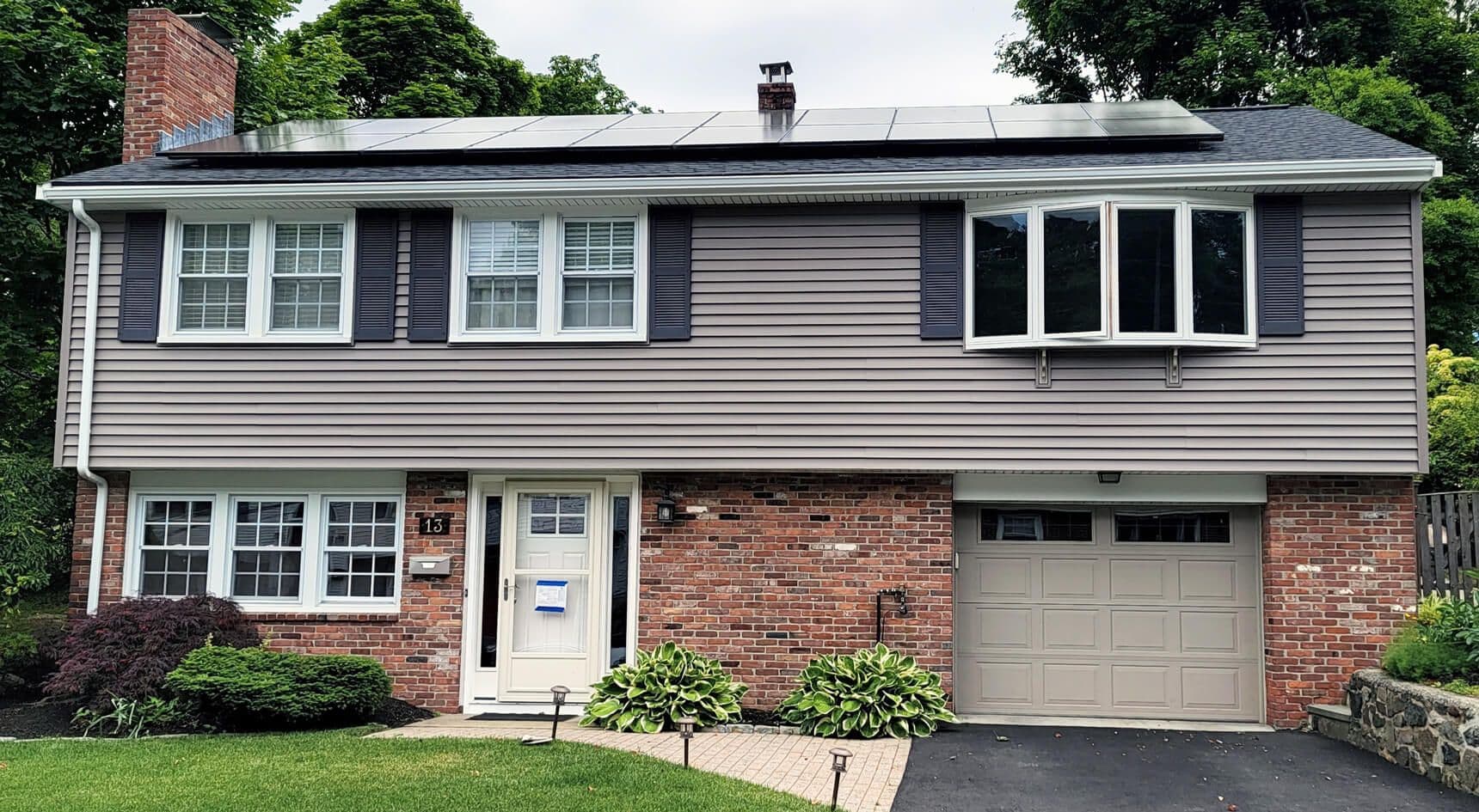 A two-story suburban house with brick and siding exterior. Solar panels line the roof. The front yard features trimmed bushes and a paved walkway.