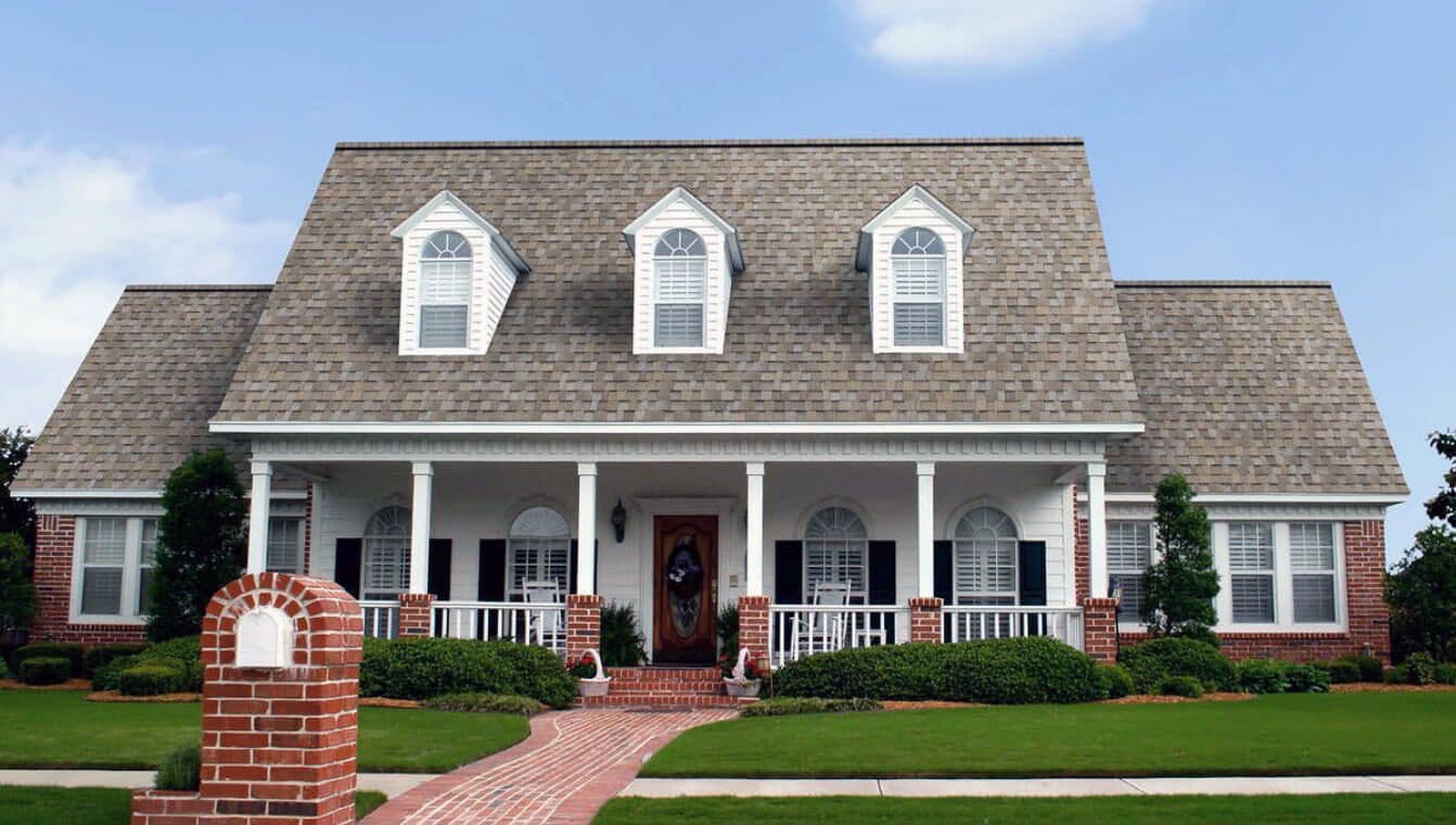 A classic brick house with a large shingled roof, three dormer windows, and white columns on the porch. Surrounded by manicured lawns and shrubs.