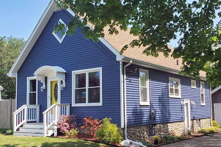 A charming blue house with white trim and a gabled roof. Lush greenery and colorful flowers adorn the front, creating a welcoming, cheerful atmosphere.