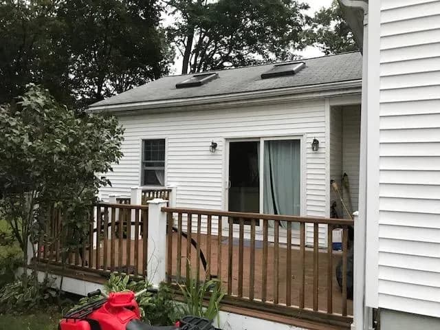 A small white house with a wooden deck, sliding glass door, and skylights on the roof, surrounded by trees. A calm, outdoor setting.