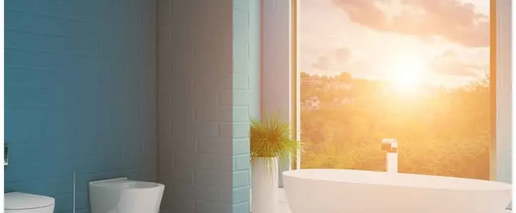 Modern bathroom with a large window showcasing a sunrise over lush greenery. Features a freestanding bathtub, toilet, and light blue walls. Calm and serene.