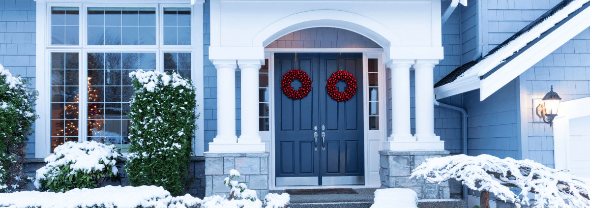 Front porch of a snowy house with two red wreaths on blue double doors. Lit Christmas tree visible through window, creating a festive winter scene.