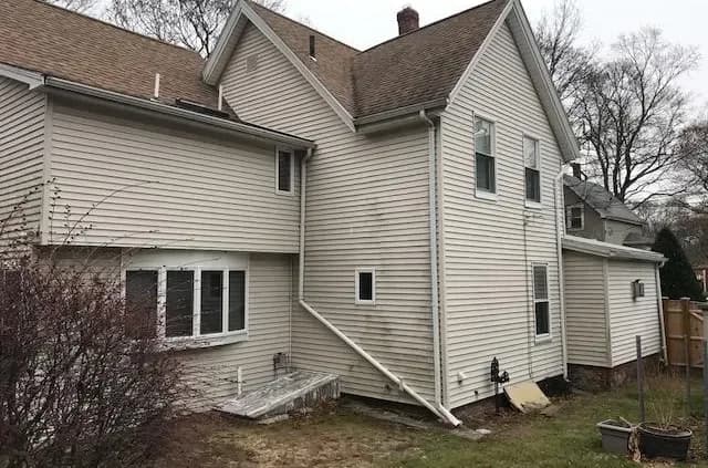 Two-story house with light beige siding, multiple gables, and a sloped roof. The yard is unkempt, featuring bare patches and leafless shrubs. Overcast sky.