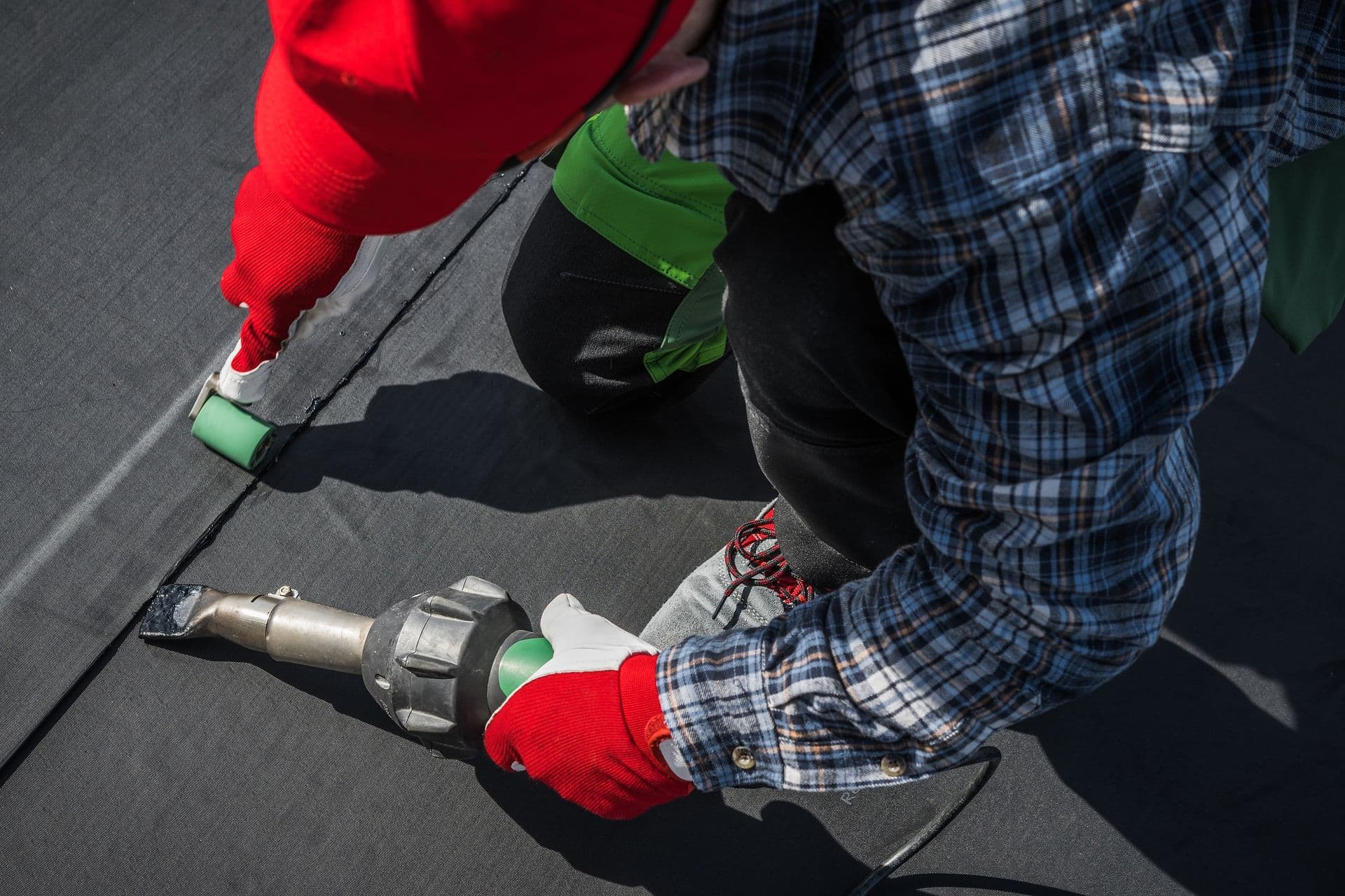 A person in a red hat and gloves uses a heat gun to seal black roofing material. They are wearing a plaid shirt and green pants, focused on precision.