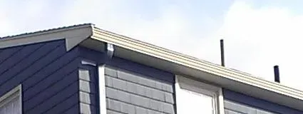 A blue house roof with white trim under a partly cloudy sky. The siding is horizontal and there are two vertical black exhaust pipes on the roof.