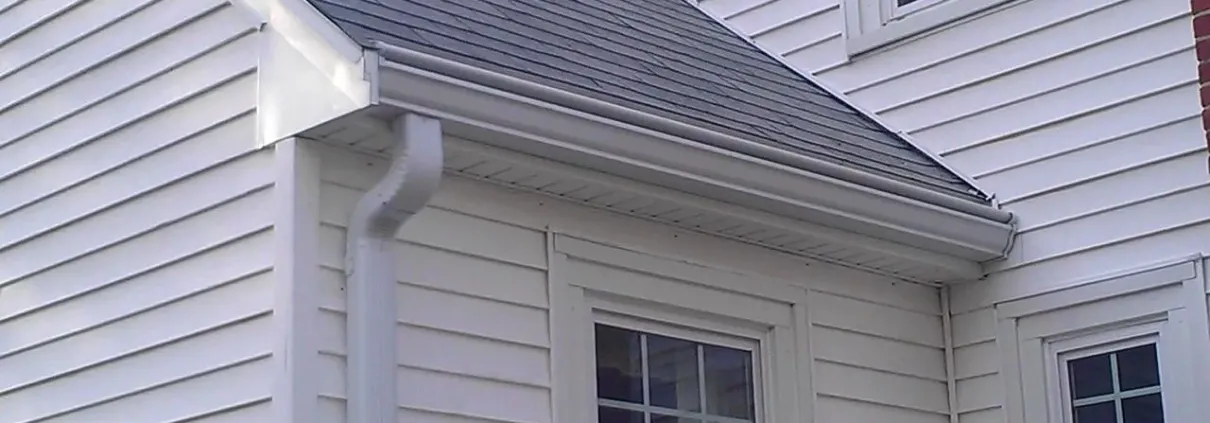 White vinyl siding house corner with two windows. Roof covered with dark shingles, and a white downspout on the left. Bright, clean exterior.