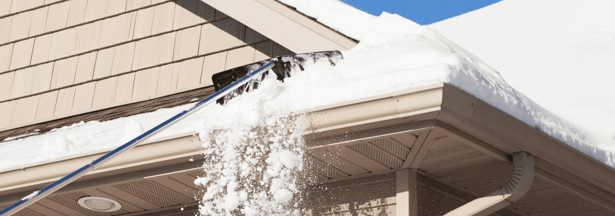 A snow-covered roof with shingles is being cleared by a rake. Snow falls off the roof's edge, contrasting against a bright blue sky.