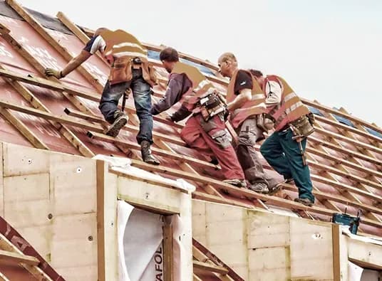 Four workers in safety vests install roof tiles on a sloped roof, displaying teamwork and focus. The scene conveys collaboration and diligence.