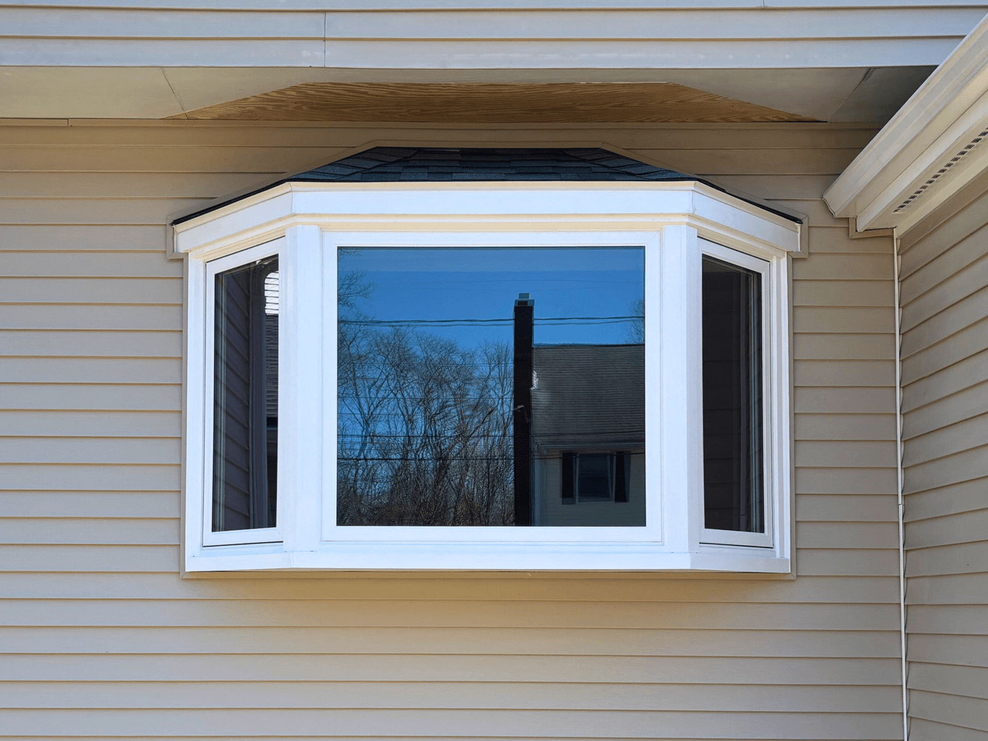 A white bay window extends from a beige house exterior with siding. Reflections of trees and a neighboring house are visible on the glass under a clear sky.