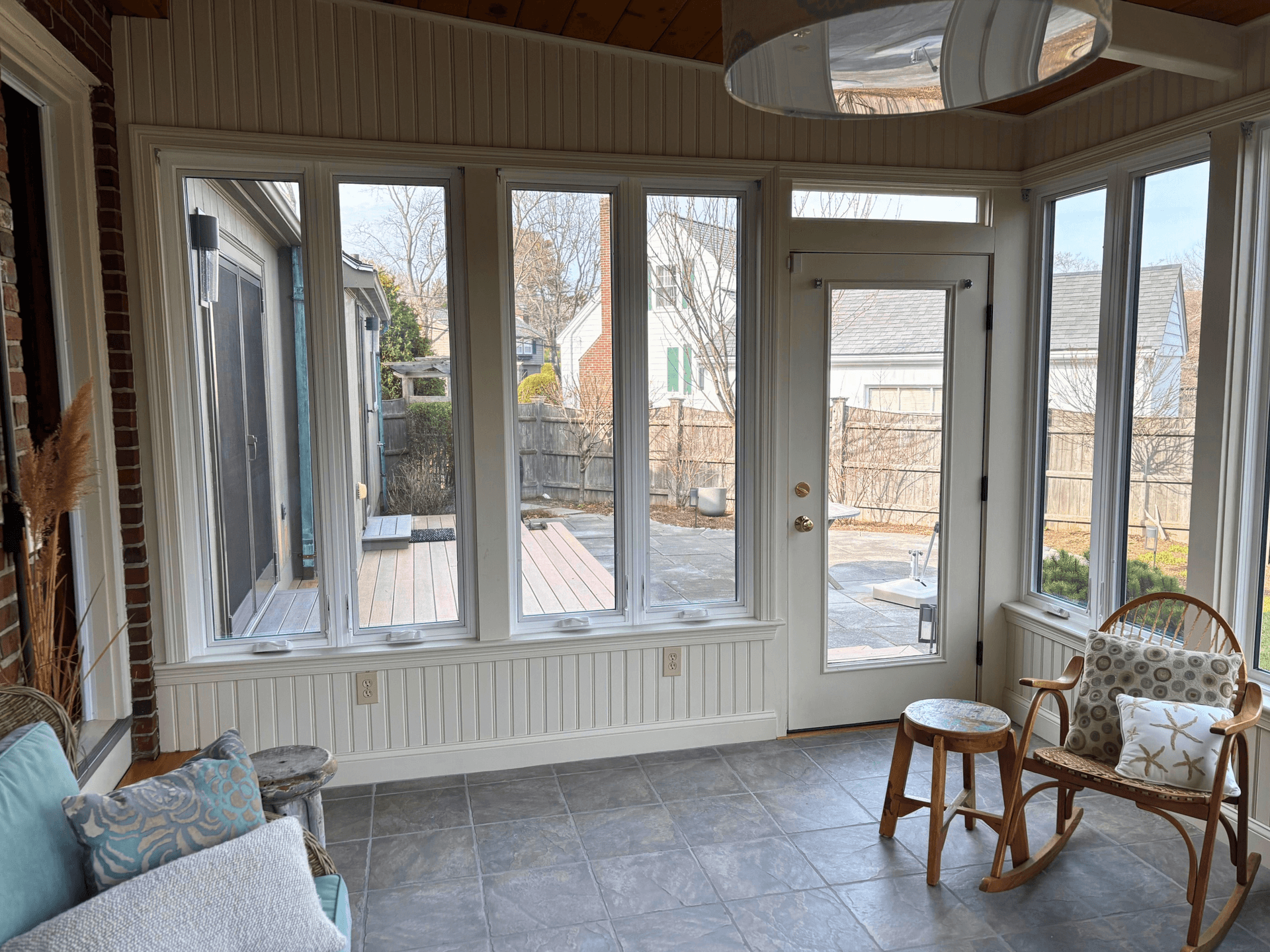 A bright sunroom with large windows overlooking a fenced backyard. Cozy wicker chair with patterned pillows, tile floor, and light, airy atmosphere.