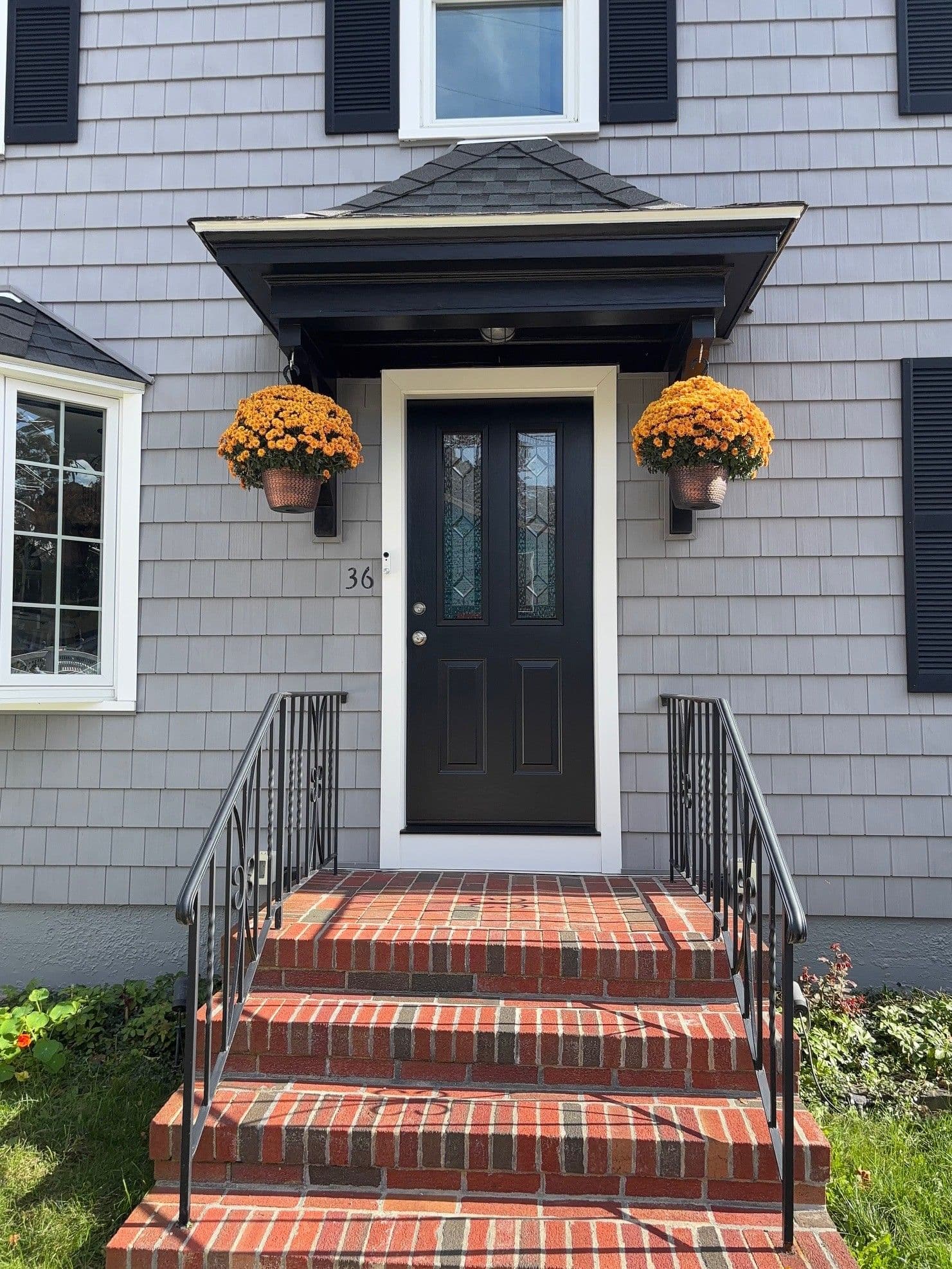 A charming front entrance with a black door, flanked by two hanging baskets of bright orange flowers. Red brick steps lead up to the door, under a gabled awning.