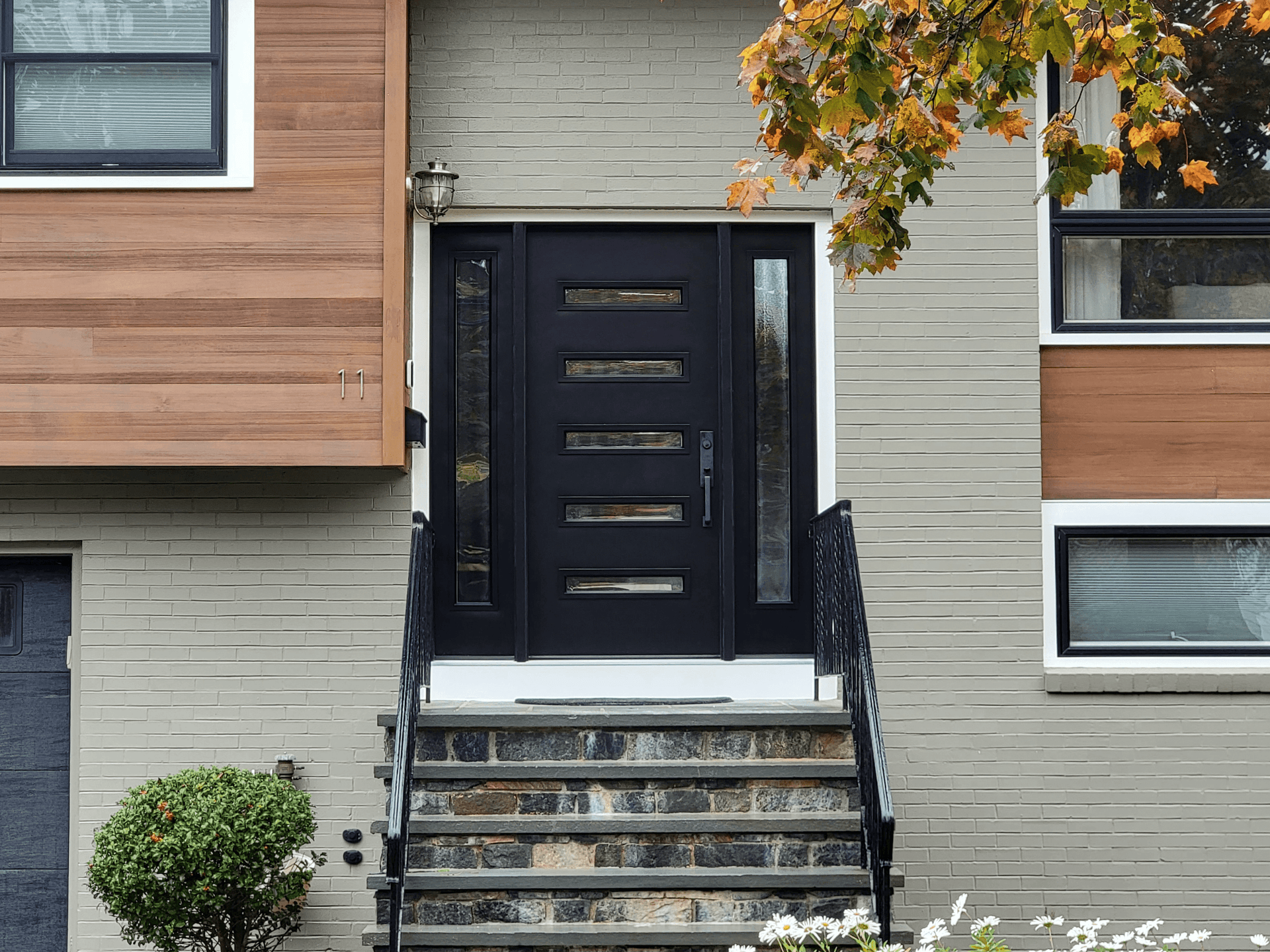 Modern front entrance with a sleek black door featuring horizontal glass panels. Stone steps lead up, flanked by a small shrub. Autumn leaves hang above.