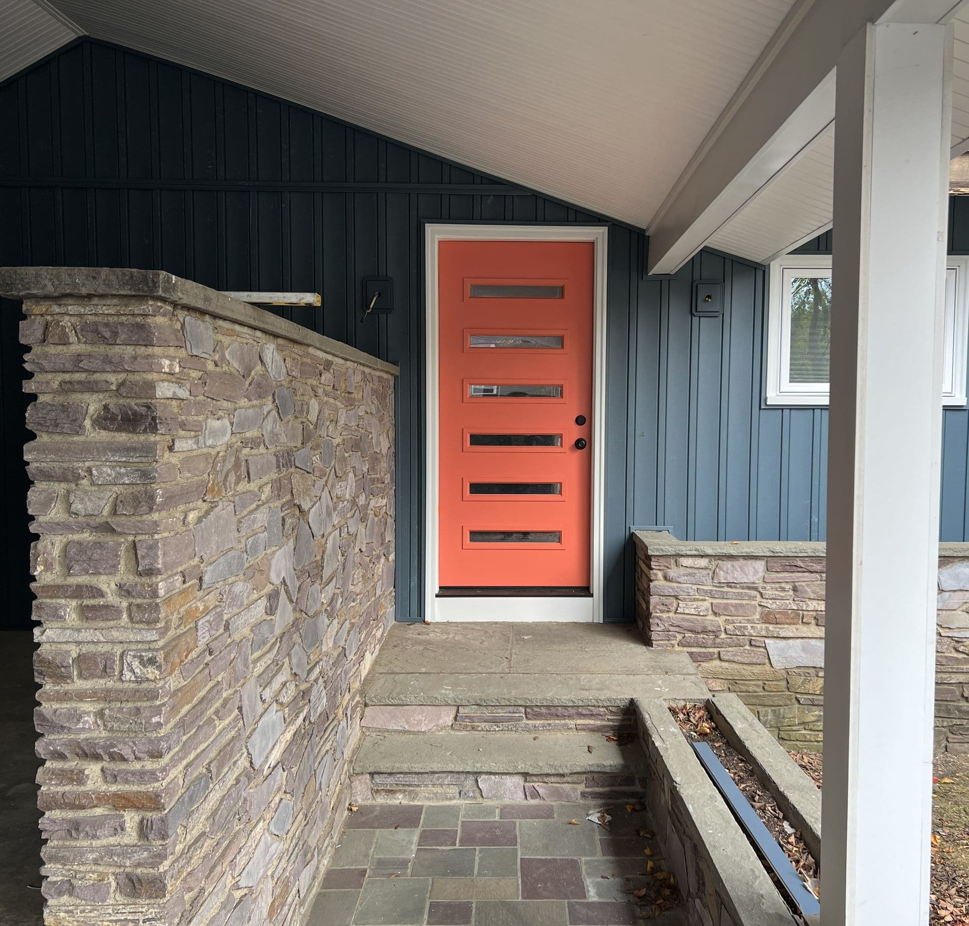 Mid-century modern porch featuring a bright coral door with rectangular windows, surrounded by blue siding and stone wall accents. Calm, inviting atmosphere.